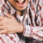 Hombre sosteniendo su hombro con expresión de dolor, vestido con una camisa de cuadros. La imagen ilustra el dolor crónico en las articulaciones.